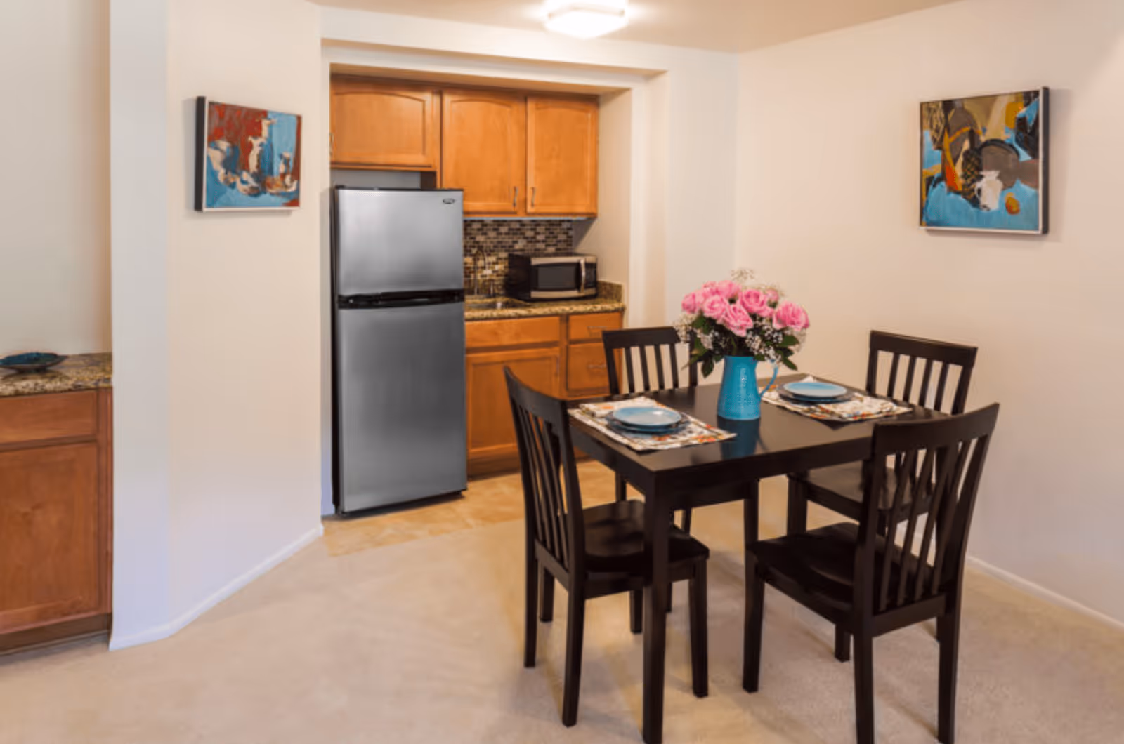 A small dining area with a dark wooden table set for two, featuring blue plates and floral placemats. A turquoise vase with pink flowers is centered on the table. Behind the dining area is a compact kitchen with wooden cabinets, a stainless steel refrigerator, a microwave, and a tiled backsplash. Two colorful abstract paintings hang on the walls.