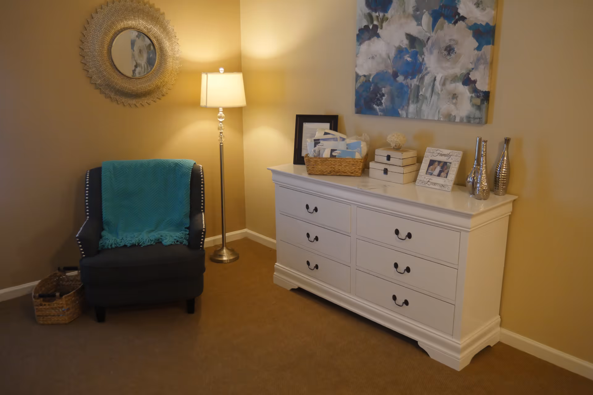 Cozy interior with an upholstered armchair and floor lamp beside a white dresser topped with decorative items and a floral painting.