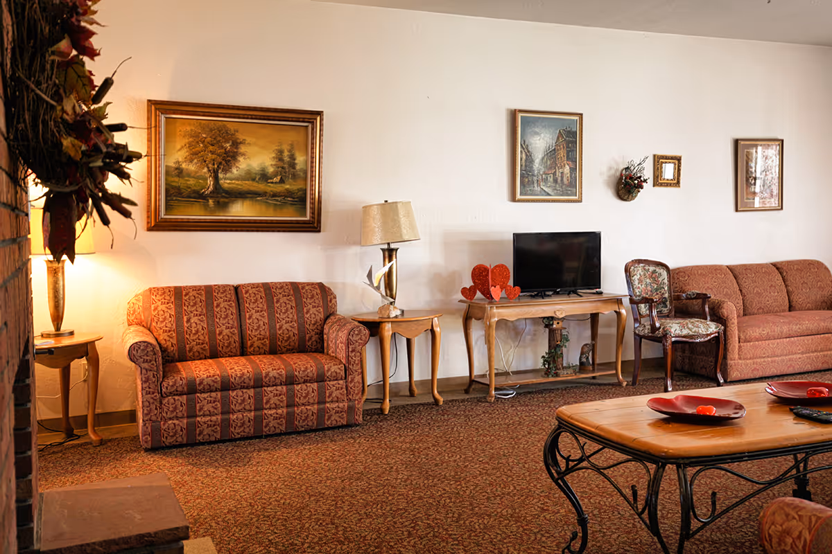 A cozy living room with two patterned sofas, a wooden coffee table with red decorative plates, a small TV on a wooden table, a floral upholstered chair, and framed paintings on the wall. There is a lamp on a side table and a fireplace partially visible on the left.
