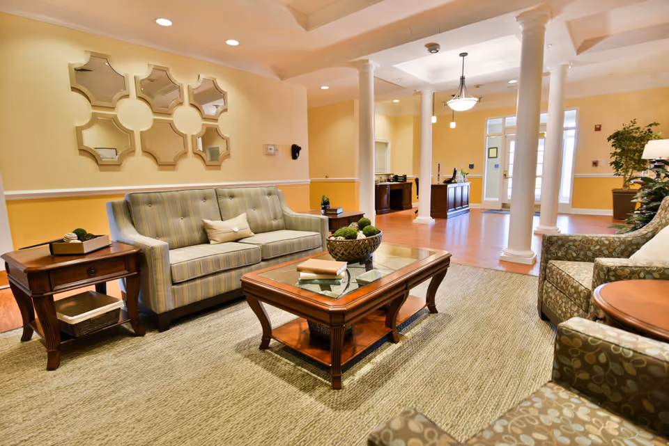 A well-lit living room area with a striped sofa, two patterned armchairs, a wooden coffee table with books and decorative items, and a side table with a small plant. The room features beige walls, white columns, a large mirror with geometric shapes on the wall, and hardwood flooring with a large area rug.