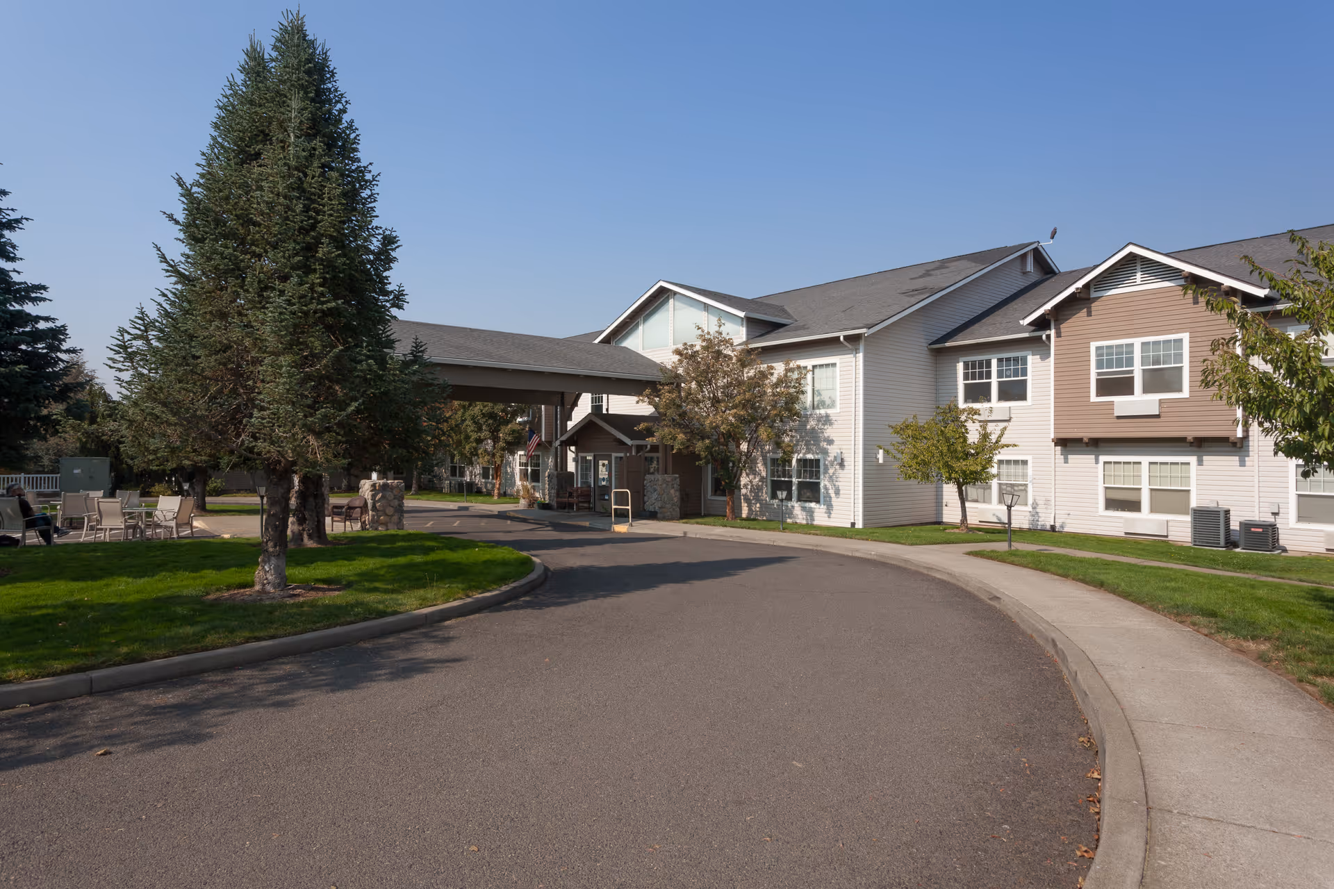 Exterior view of Skylark Assisted Living facility showing a two-story building with a covered entrance, surrounded by trees, green lawns, and a curved driveway under a clear blue sky.