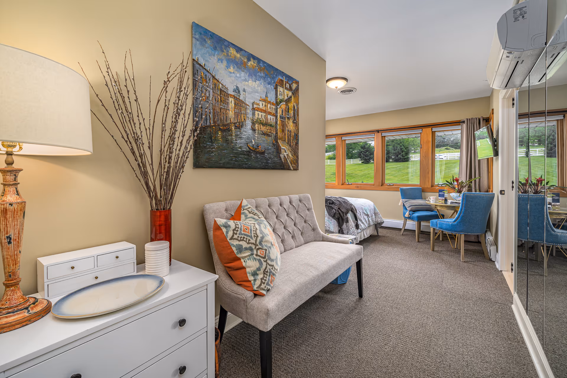 A cozy senior living room area with a beige tufted loveseat adorned with patterned pillows, a white chest of drawers with a decorative lamp and vase with branches, a large painting of a canal scene on the wall, and a windowed bedroom area in the background featuring a bed, blue chairs, and a small table with plants and cups. The room has carpeted flooring and a mirrored closet door on the right.