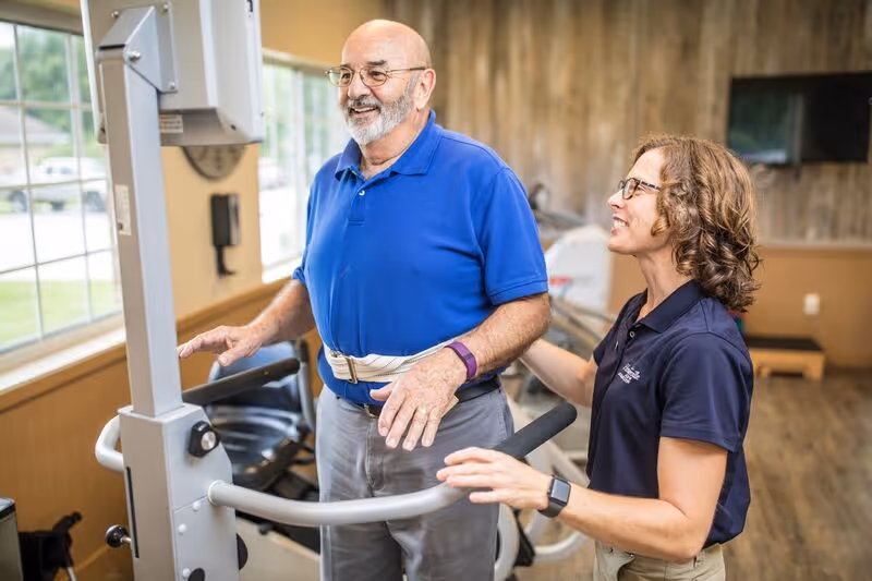 An elderly man wearing a blue polo shirt and gray pants is using a piece of exercise equipment in a well-lit room with large windows. A woman in a navy blue polo shirt, possibly a staff member, is standing beside him, providing support and encouragement. The room has wooden paneling on the walls and exercise machines in the background.