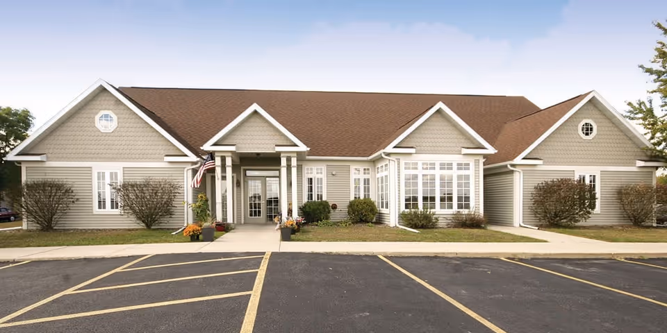 Front exterior view of a single-story senior living facility building with beige siding, a brown roof, multiple windows, an American flag near the entrance, and a parking lot in front.