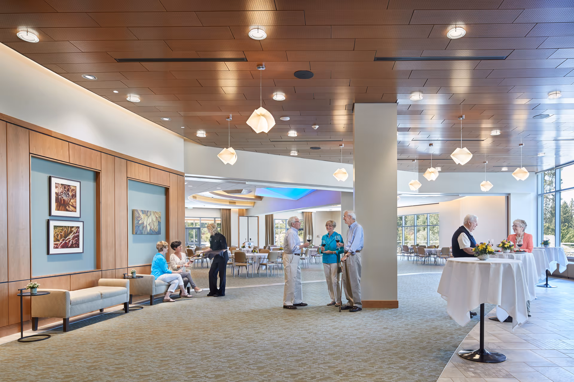 A spacious, well-lit common area in a senior living facility with several elderly people engaged in conversation. The room features modern ceiling lights, large windows letting in natural light, and tables with white tablecloths and floral centerpieces. There are also comfortable seating areas along the wooden paneled wall with framed artwork.