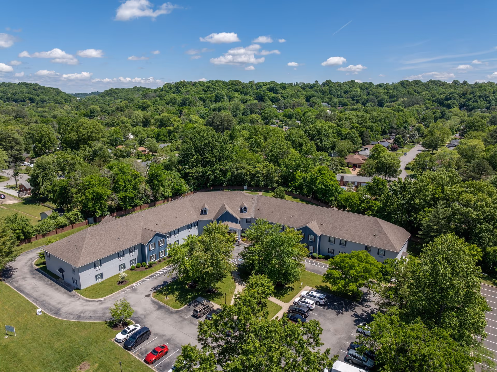 Aerial view of The Village at Bellevue, a large assisted living facility surrounded by lush green trees and residential neighborhoods under a blue sky with scattered clouds. The building has a brown roof and light-colored exterior walls, with a parking lot containing several cars in front.