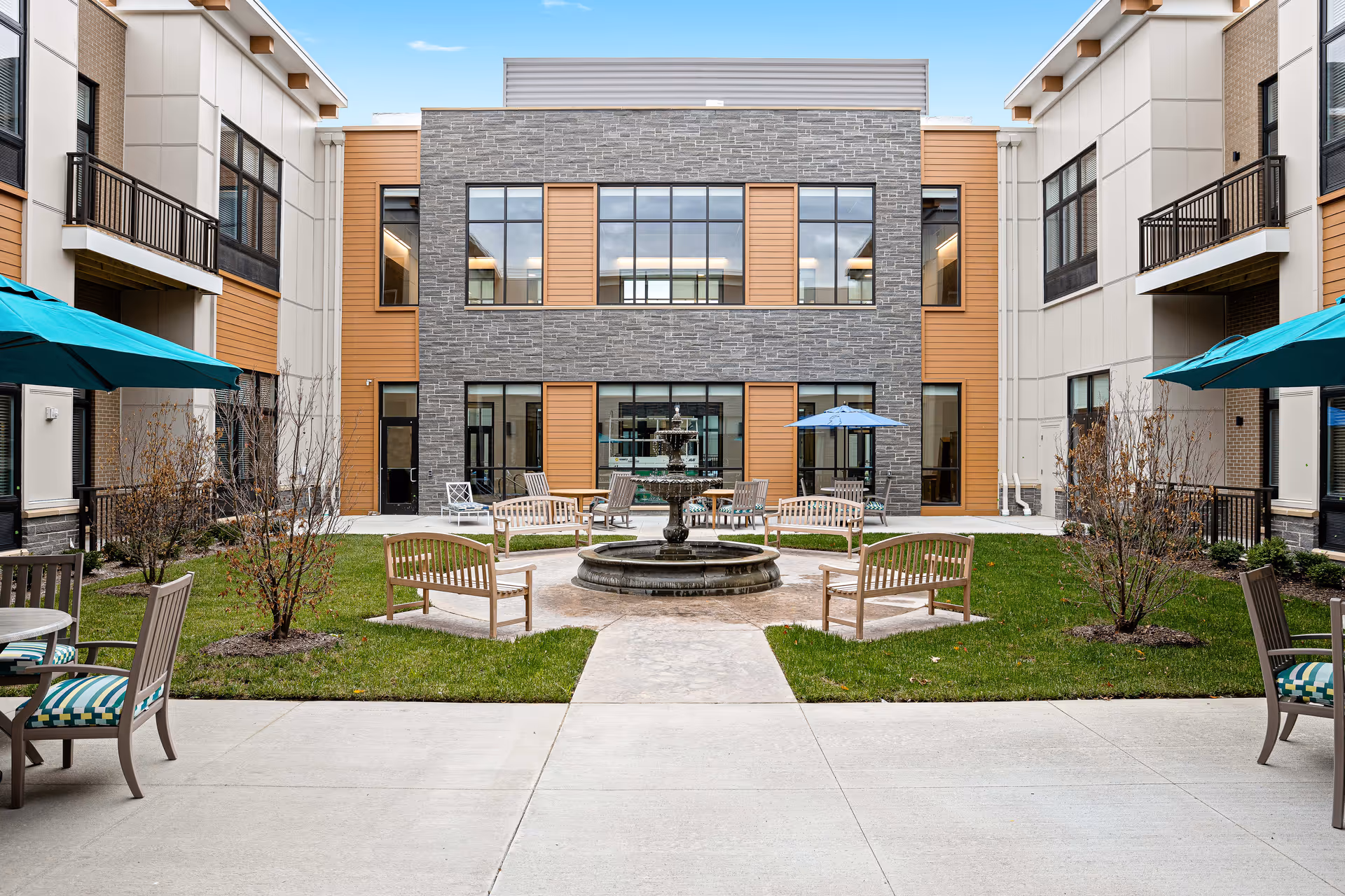 Courtyard at a senior living facility with benches and a central fountain surrounded by patio tables, umbrellas, and a two-story building.