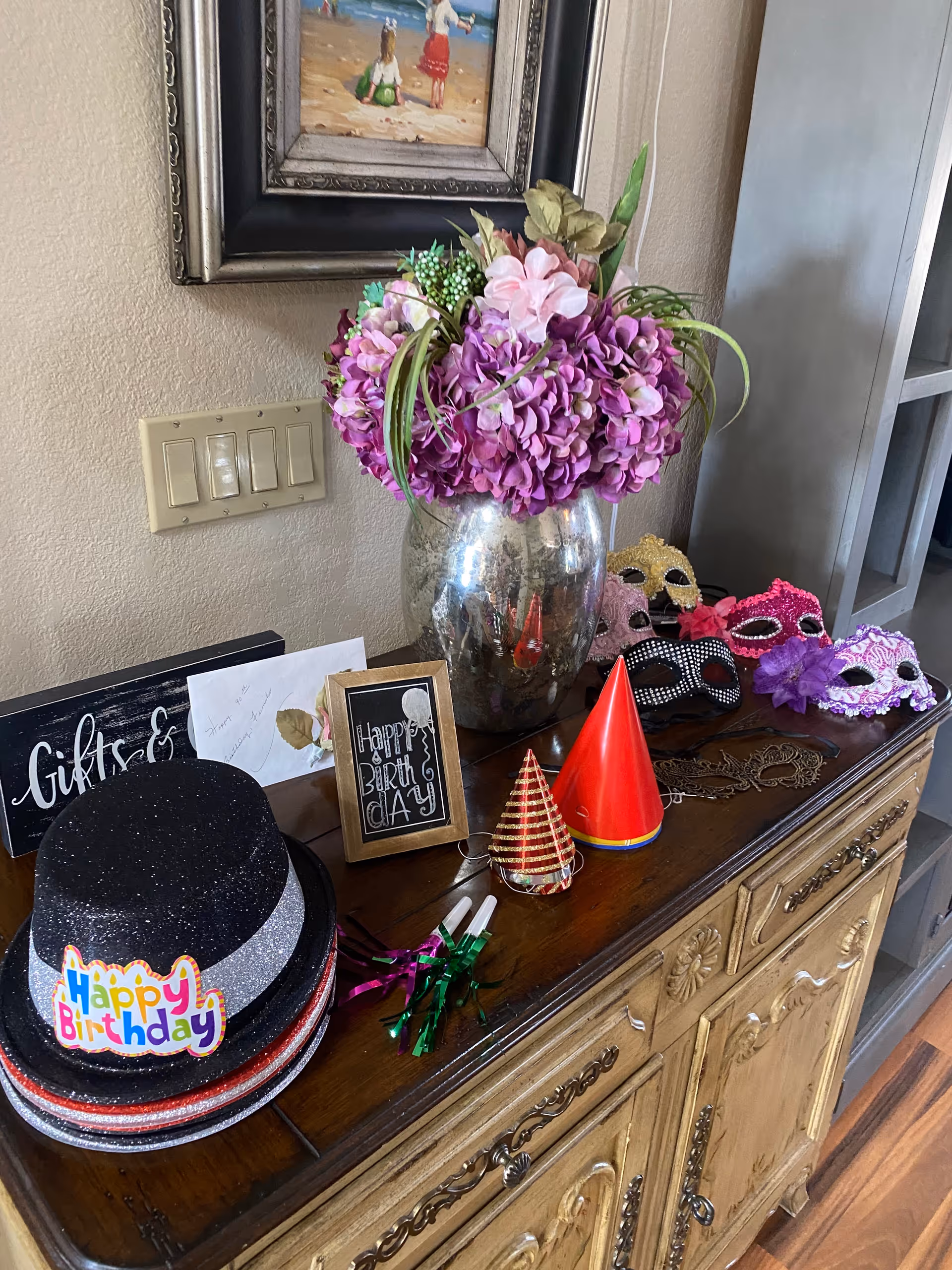 A wooden sideboard decorated with birthday party items including a black glittery top hat with a 'Happy Birthday' sticker, colorful party hats, noisemakers, and decorative masks. A large silver vase with purple and pink flowers sits on the sideboard. On the wall above, there is a light switch panel and a framed painting of two children on a beach.