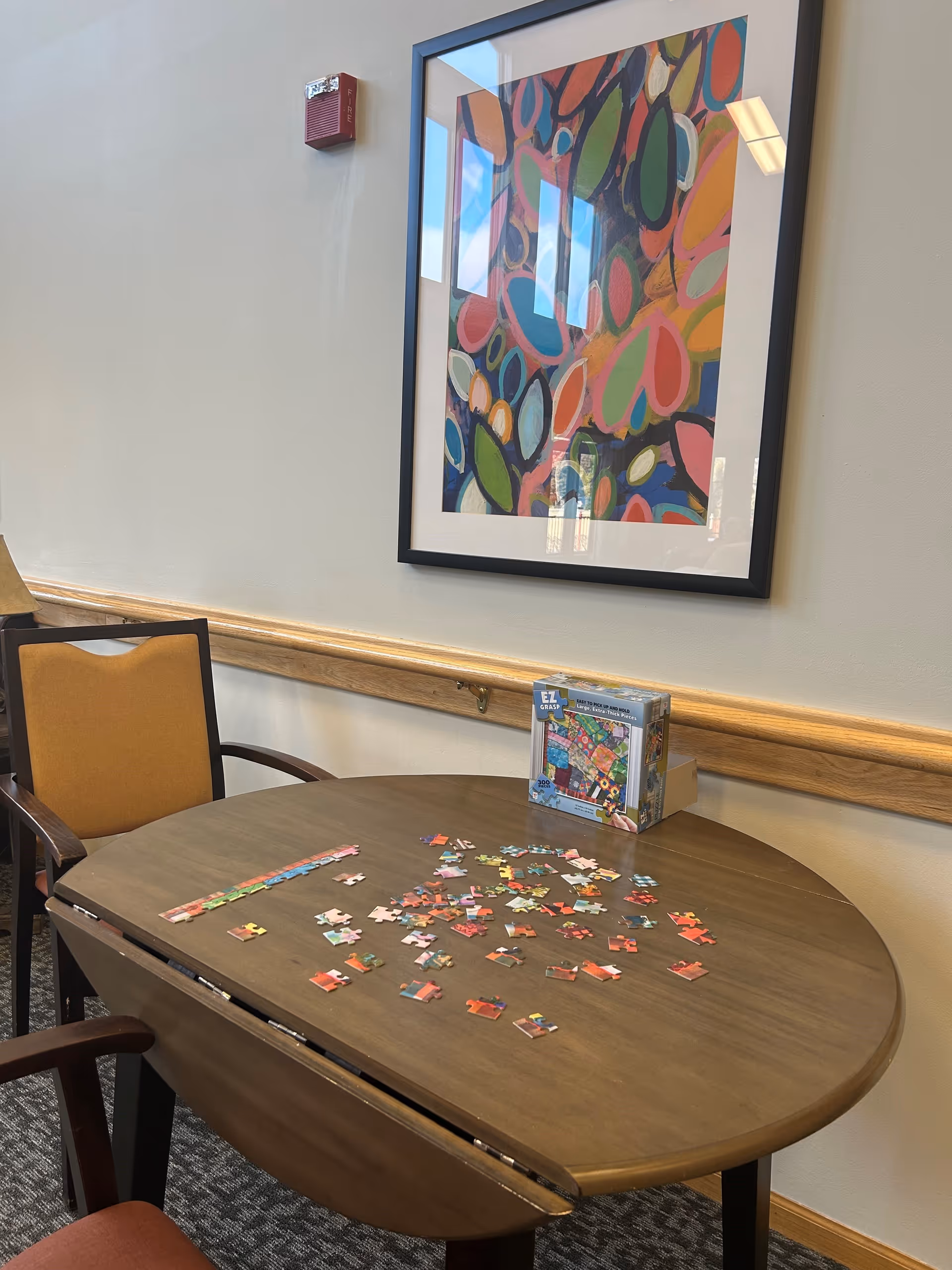 A round wooden table with scattered puzzle pieces and a puzzle box on it, surrounded by chairs with orange cushions. A colorful abstract painting is hung on the wall above the table, and a fire alarm is mounted on the wall.