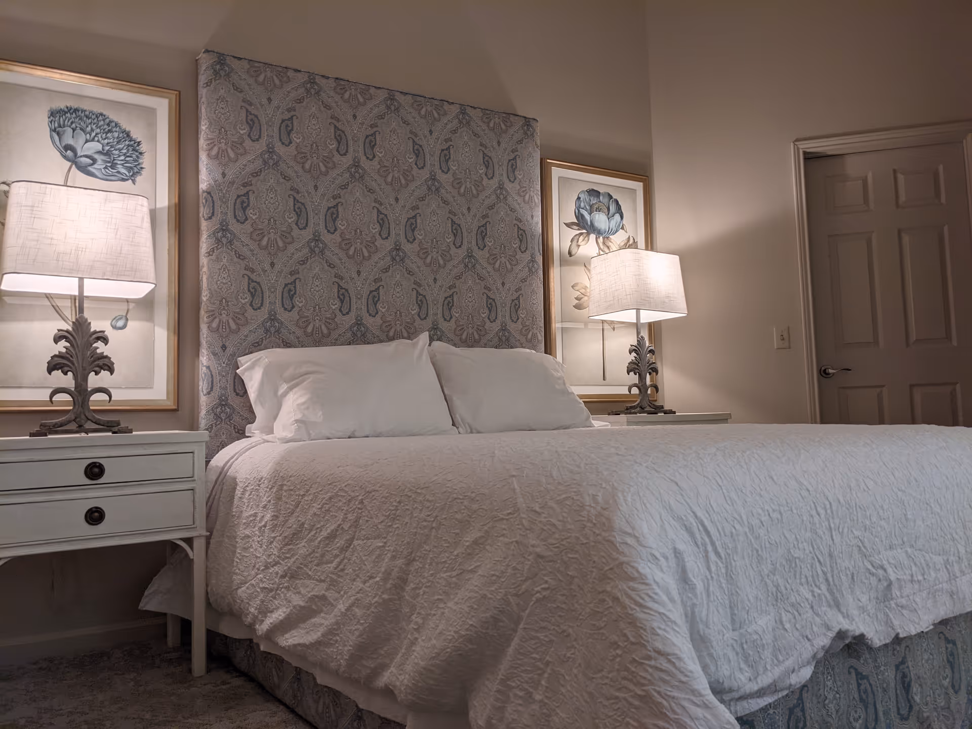 A bedroom with a large bed covered in a white quilt and two white pillows. The bed has a tall, patterned headboard. On each side of the bed, there is a white nightstand with two drawers and a decorative lamp with a white shade. Behind each lamp, there is framed artwork featuring blue flowers. A closed door is visible on the right side of the room.