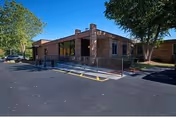 Exterior view of a single-story brick building with large windows and a ramp leading to the entrance, surrounded by trees and a paved parking area.