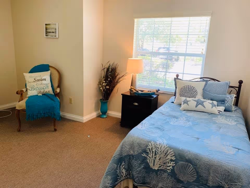A cozy bedroom with a single bed covered in blue bedding featuring sea-themed patterns. The bed has several decorative pillows with starfish and seashell designs. Next to the bed is a black nightstand with a lamp and decorative items. A window with blinds lets in natural light. To the left, there is a wooden chair with a blue throw blanket and a pillow with beach-themed words. A small framed picture hangs on the wall above the chair.