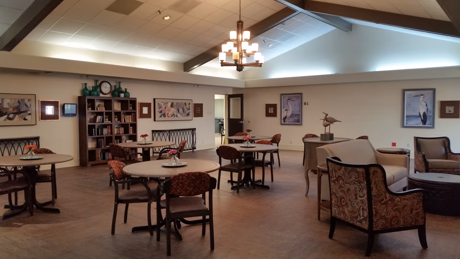Large communal sitting and dining room with round tables, chairs, bookshelves and lounge seating under a vaulted ceiling.