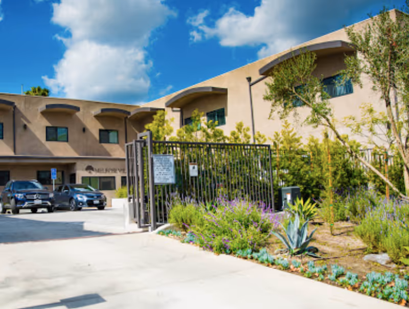 Exterior view of Melrose Villas senior living facility showing a two-story beige building with several windows, a gated entrance, parked cars, and landscaped greenery including shrubs and small trees under a partly cloudy blue sky.