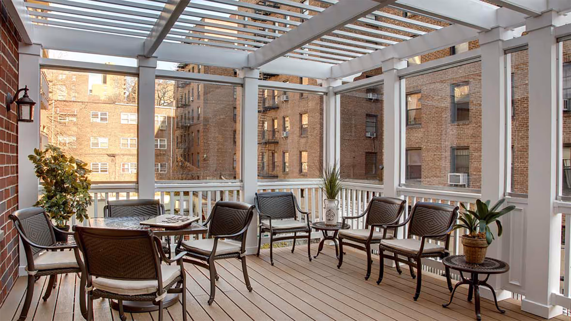 A covered outdoor patio area with a wooden floor, white beams overhead, and glass panels enclosing the space. The patio is furnished with several cushioned chairs arranged around a round table and two small side tables with potted plants. Brick buildings are visible outside the glass panels.