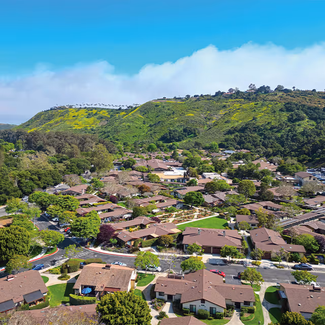 Aerial view of a senior living community named Valle Verde, showing multiple single-story residential buildings with brown roofs surrounded by trees and greenery. In the background, there are green hills under a clear blue sky.