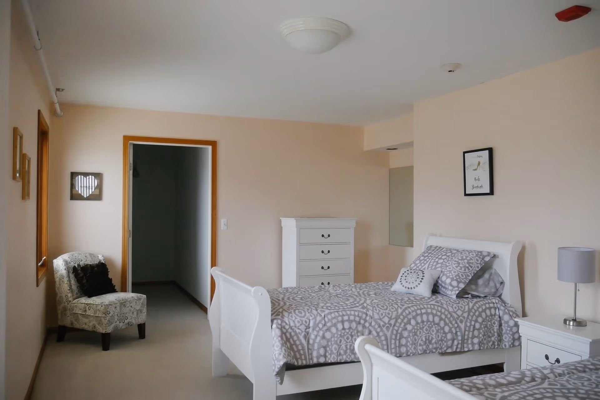 A senior living bedroom with two white wooden beds featuring gray and white patterned bedding. There is a white chest of drawers against the far wall, a small white nightstand with a gray lamp, and a patterned armchair with a dark cushion near a window. The walls are light beige with minimal decorations, including a framed picture and a heart-shaped wall hanging.