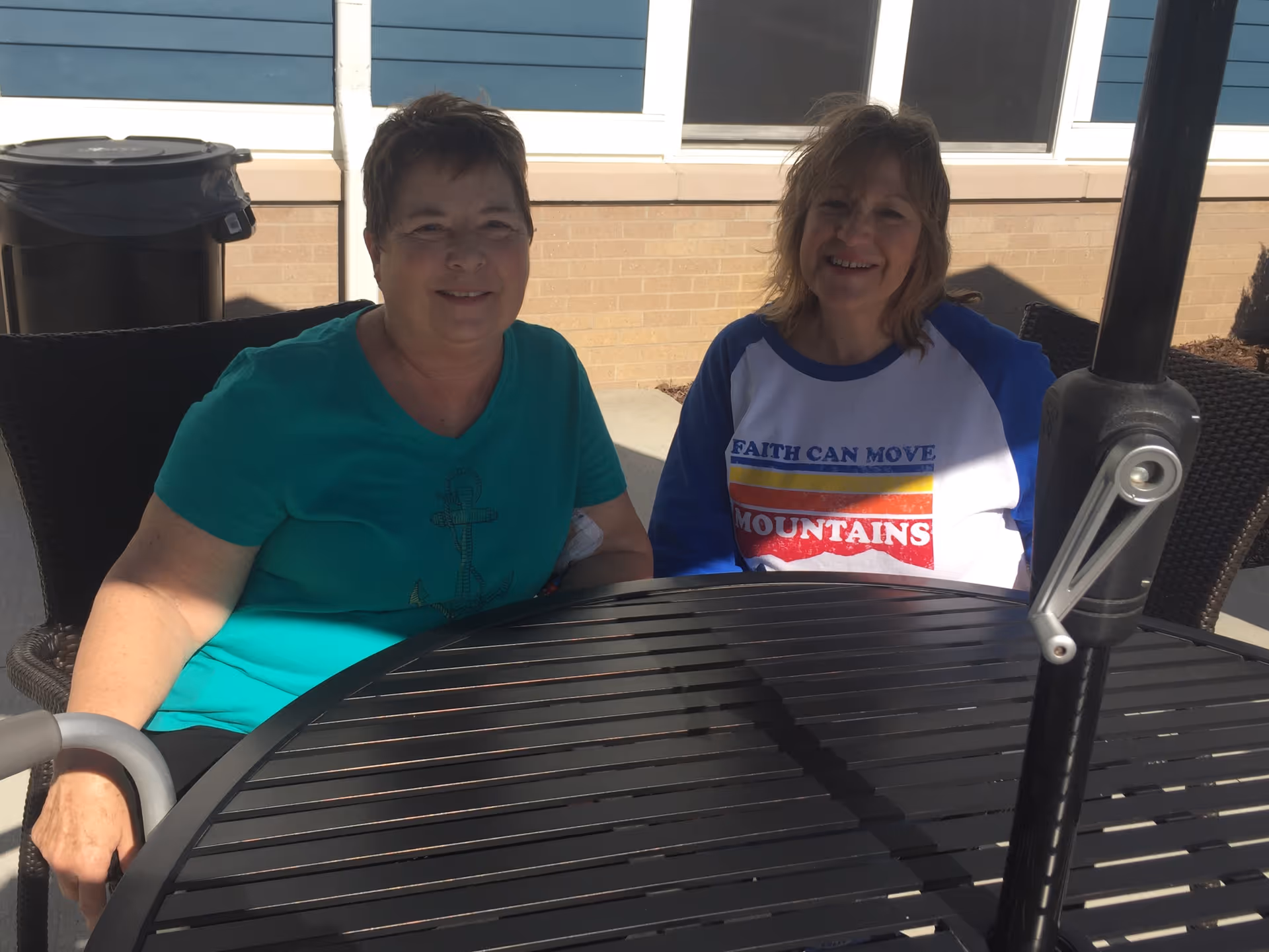 Two women sitting at a round outdoor table with a black umbrella pole in the center. One woman is wearing a teal shirt with an anchor design, and the other is wearing a white and blue shirt with the text 'Faith Can Move Mountains'. They are seated outside near a building with blue siding and beige brick.