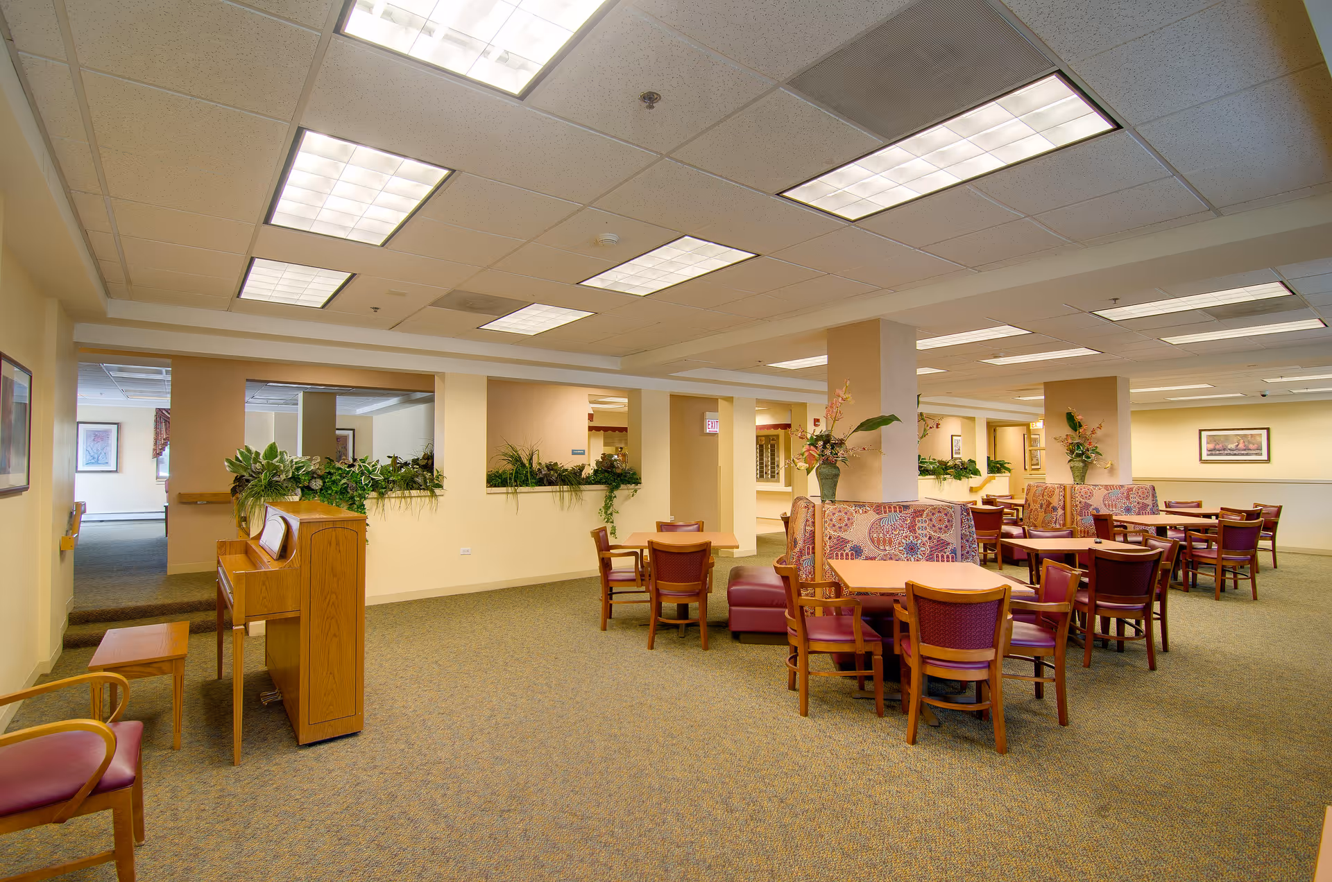 Interior view of a senior living facility common area with multiple wooden tables and chairs arranged for dining or socializing. There are patterned cushioned booth seats around some tables, potted plants on pillars, and a wooden piano with a bench on the left side. The ceiling has recessed fluorescent lighting, and the walls are painted in light neutral colors.