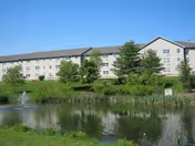 A large multi-story residential building with beige siding and multiple windows, situated behind a pond with a small fountain and surrounded by green grass and trees under a clear blue sky.
