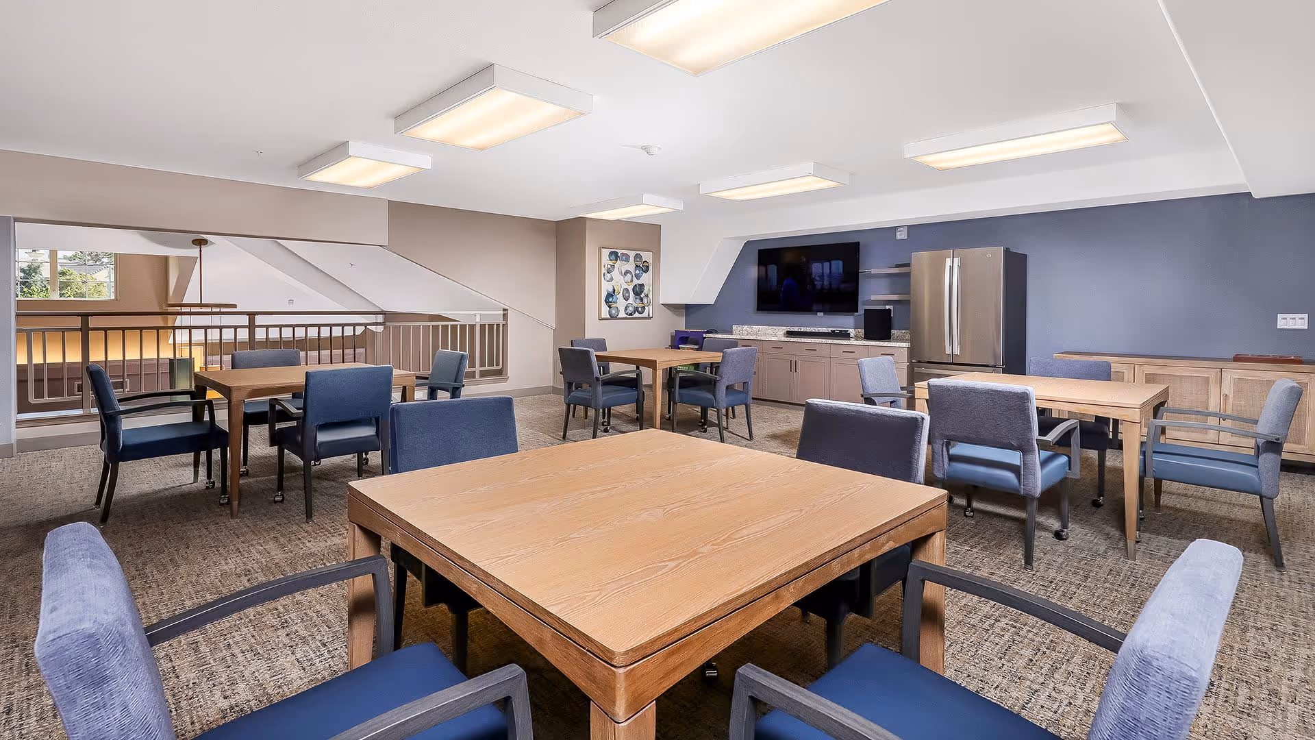A spacious common area with several wooden tables and blue cushioned chairs arranged around them. The room features a carpeted floor, a large flat-screen TV mounted on a blue accent wall, a stainless steel refrigerator, and cabinetry. Overhead fluorescent lighting illuminates the space, and there is a railing overlooking a lower level visible in the background.