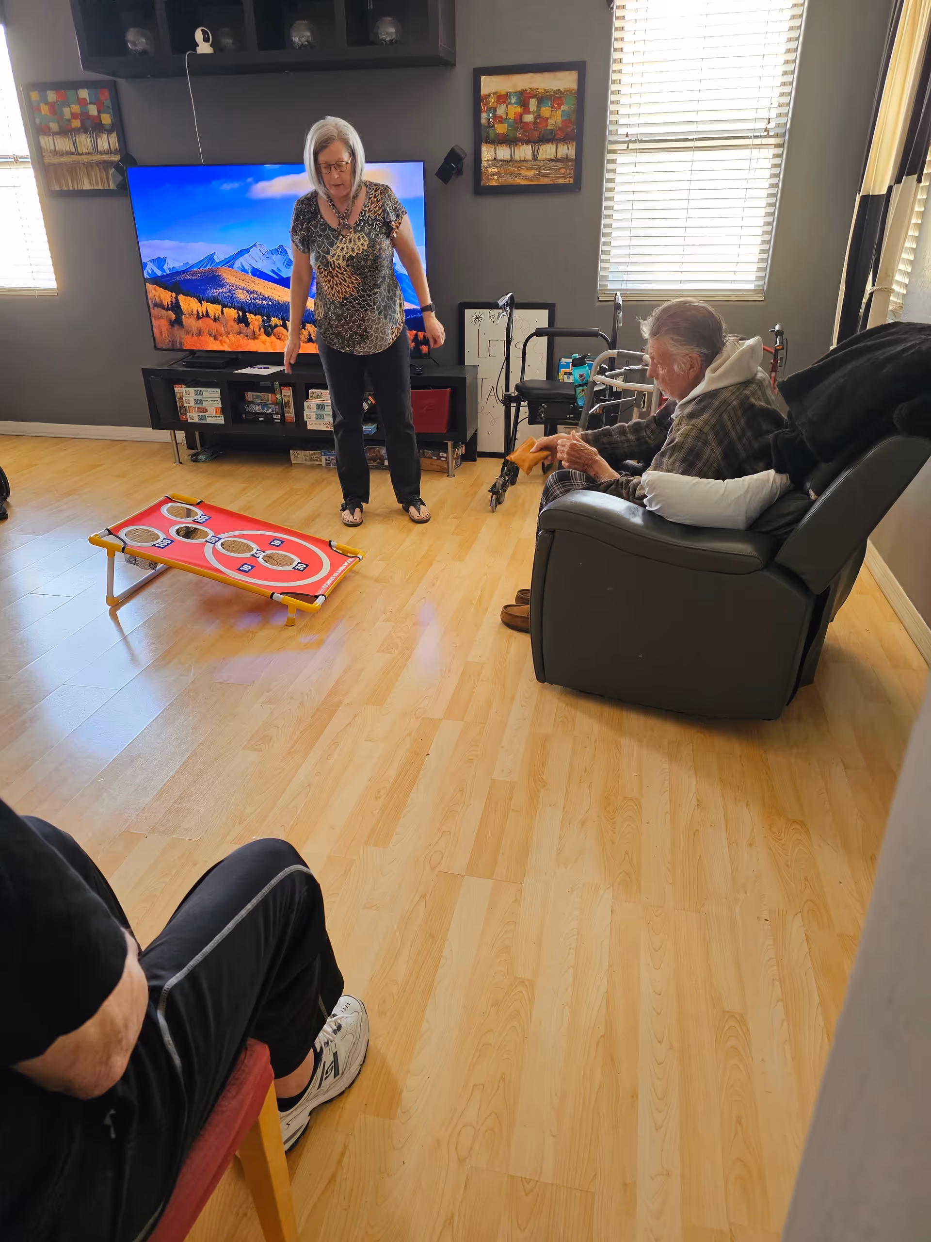 A senior woman standing and playing a bean bag toss game in a living room with two other seniors seated, one in a recliner and the other on a chair. The room has wooden flooring, a large TV on a stand, colorful paintings on the walls, and windows with blinds letting in natural light.
