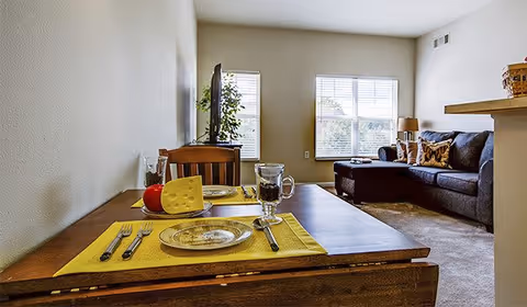 A cozy living room and dining area with a wooden table set for two with yellow placemats, plates, cutlery, a glass pitcher, and a decorative cheese and apple. In the background, there is a dark gray sectional sofa with pillows, a lamp on a side table, and two windows letting in natural light.