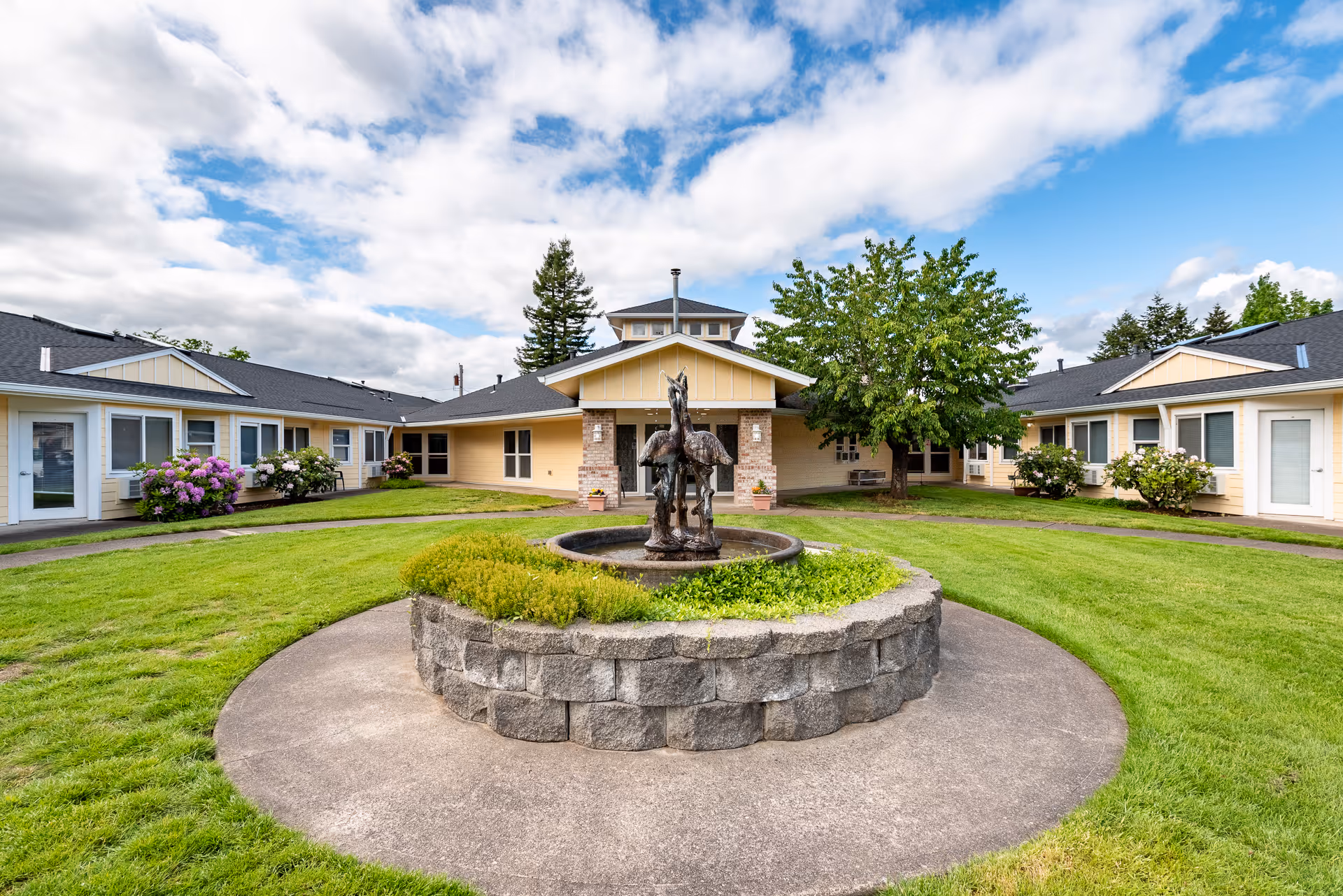 Courtyard with a circular stone fountain and horse statue in front of a single-story assisted living building surrounded by lawn and walkways.