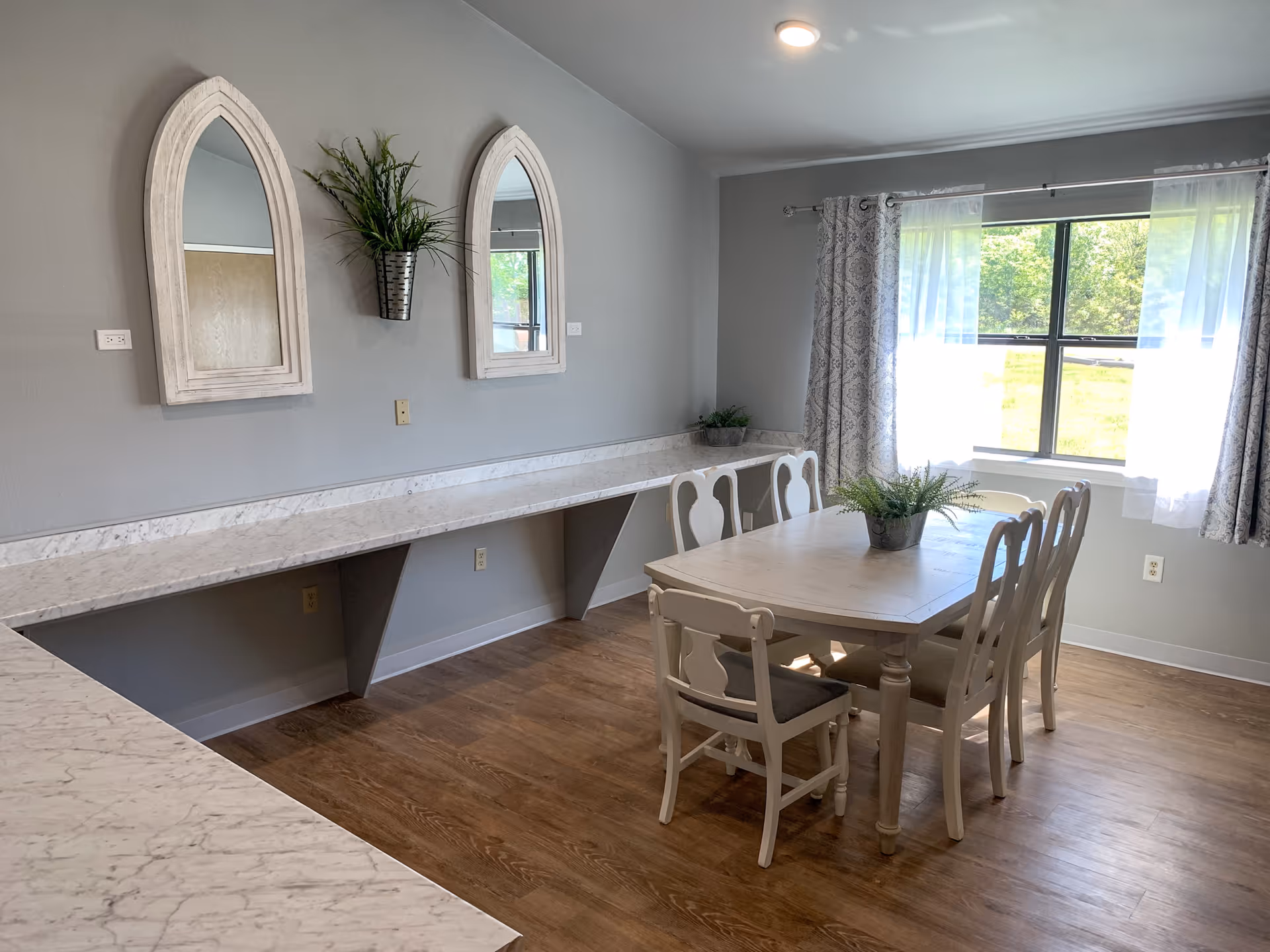 A bright dining room with a wooden table and six chairs, a large window with sheer and patterned curtains letting in natural light, two decorative mirrors on the wall, and a long marble countertop along one side of the room with potted plants for decoration.