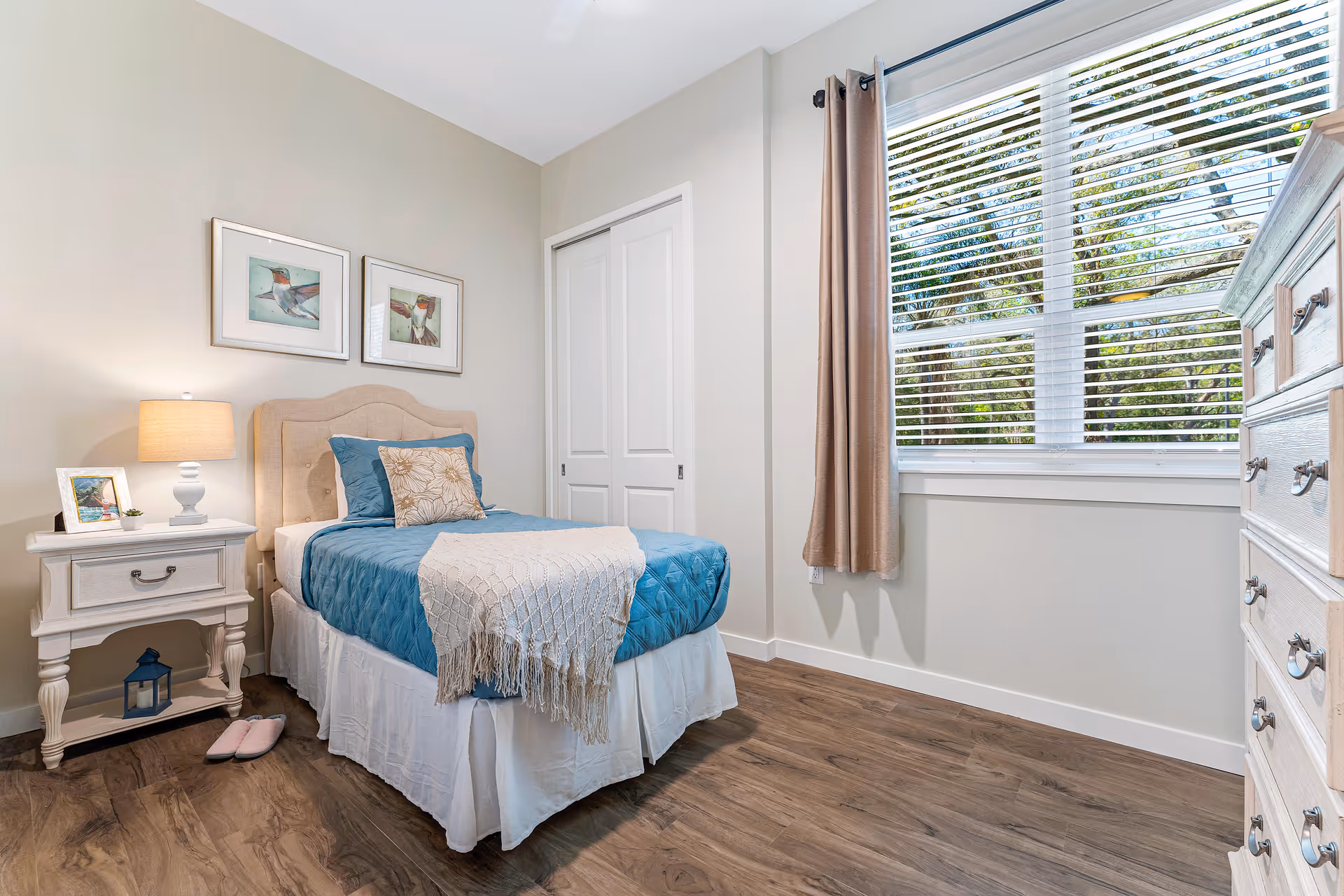 A cozy bedroom with a single bed dressed in blue bedding and a beige throw blanket. The room features a white nightstand with a lamp, a framed photo, and a small plant. Two framed bird pictures hang on the wall above the bed. There is a window with white blinds and beige curtains, and a white dresser with multiple drawers on the right side. The floor is wooden.