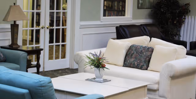 A cozy living room area with a white sofa adorned with three cushions, including one with a floral pattern. In front of the sofa is a white coffee table with a small flower arrangement. To the left, there is a blue armchair and a side table with a lamp. French doors with glass panes are visible in the background, along with a window and some greenery in the corner.