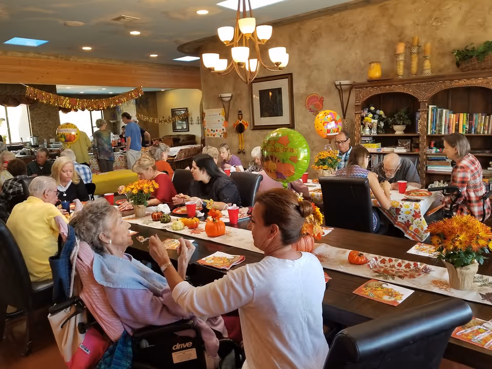A group of elderly people and caregivers gathered in a decorated dining room for a Thanksgiving celebration. The room features long tables with autumn-themed centerpieces, small pumpkins, and festive balloons. People are seated and eating, while others are standing and serving food. The walls have warm tones and shelves with books and decorations.