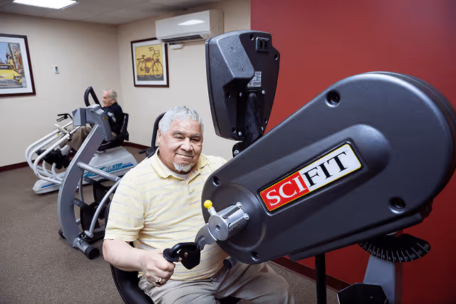 Two elderly men exercising on seated fitness machines in a room with beige walls and framed pictures, one man in the foreground smiling at the camera while using a SciFit exercise machine.