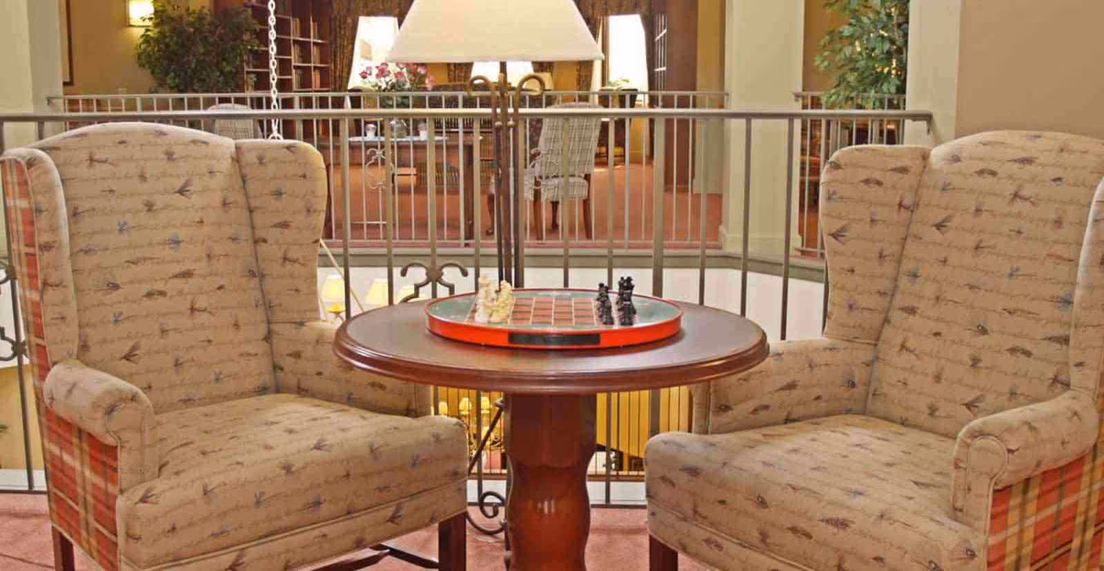Two upholstered armchairs with patterned fabric facing each other across a round wooden table with a chessboard set up on it, situated in a cozy indoor area with a railing and additional seating in the background.