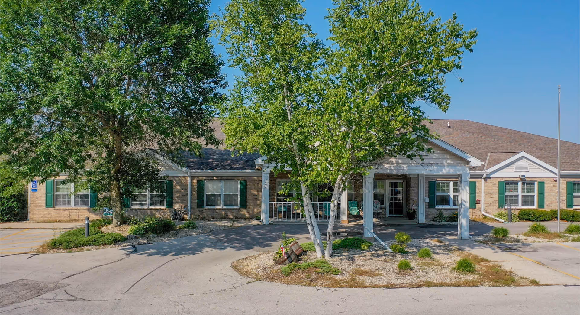 Front exterior view of Meadow View Assisted Living facility showing a single-story building with a covered entrance, green window shutters, and trees in front under a clear blue sky.