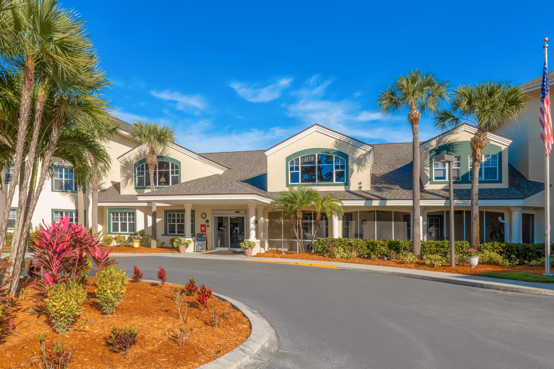 Exterior front view of Ventura Place - Sky Active Living facility with a curved driveway, palm trees, landscaped garden beds, and a clear blue sky.