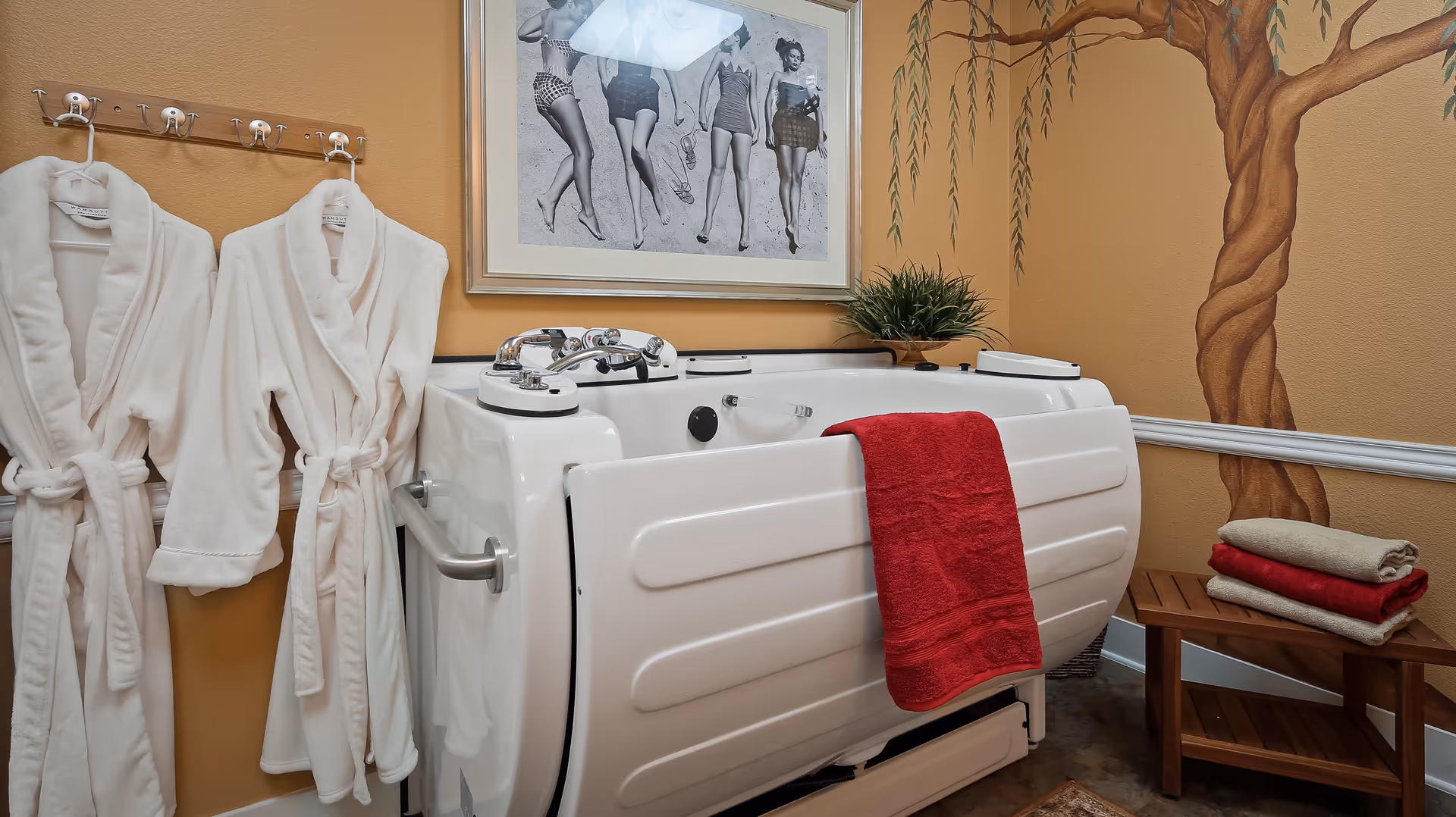 A bathroom area with a white walk-in bathtub featuring chrome fixtures and a red towel hanging on its side. Two white bathrobes hang on hooks on the left wall. A framed black and white photo of women in vintage swimsuits is mounted above the bathtub. To the right, there is a wooden bench with folded towels in red and beige. The walls are painted a warm tan color with a mural of a tree on the right side.