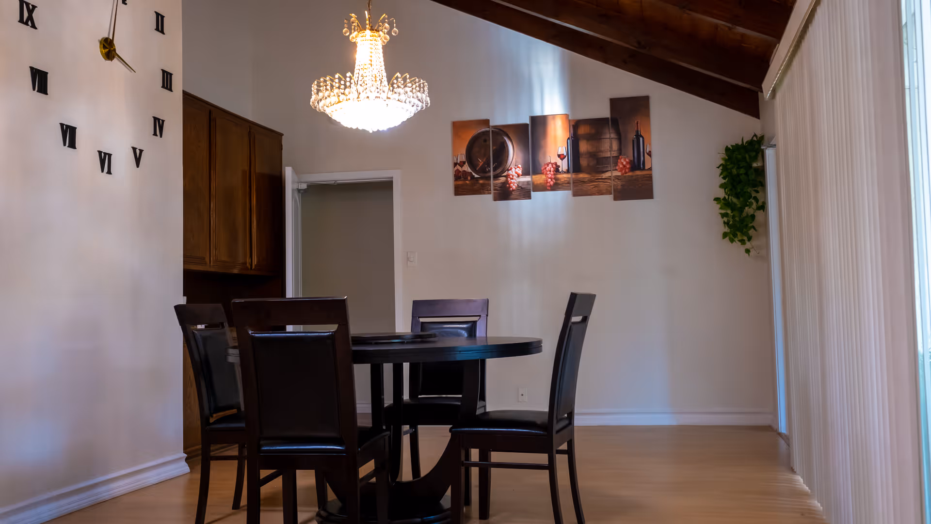 A dining room with a round dark wooden table and four matching chairs. The room features a large wall clock with Roman numerals, a crystal chandelier hanging from a wooden ceiling, a multi-panel wall art depicting wine and grapes, and a green plant in the corner near vertical blinds covering a window or door.