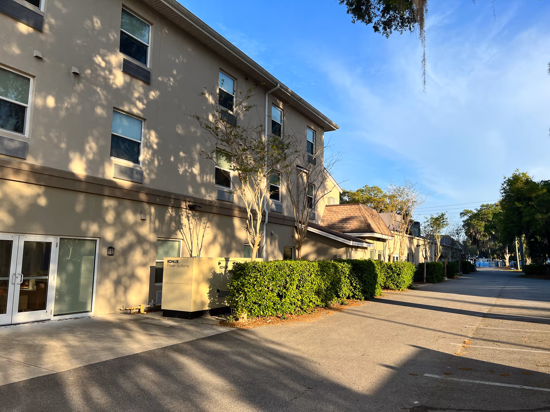 Exterior view of a multi-story senior living building with an entrance, bushes, and a parking area under a blue sky.