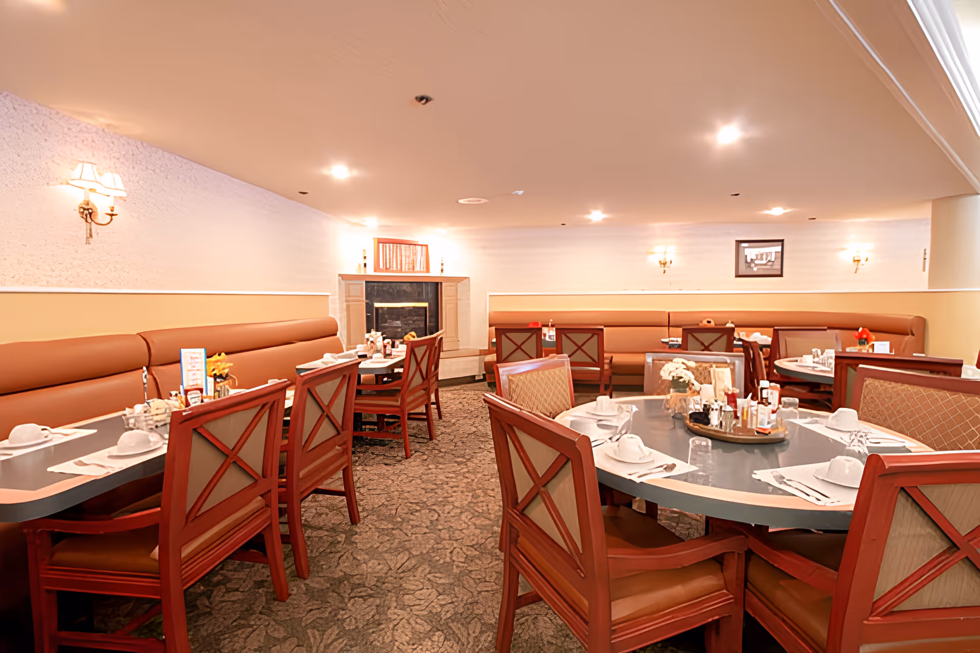A cozy dining room with multiple tables set for meals, featuring wooden chairs with cushioned seats and backs, a carpeted floor, and a fireplace in the background. The walls are light-colored with wall sconces providing warm lighting, and there is a framed picture on the far wall.