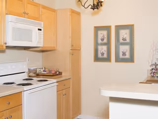 A kitchen area featuring light wood cabinets, a white microwave mounted above a white electric stove, and a white dishwasher. The countertop has a few decorative items, and two framed pictures hang on the beige wall. A chandelier is partially visible above.