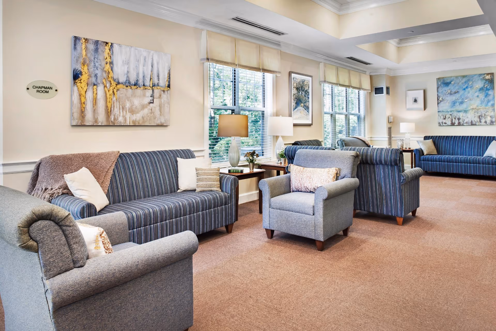 Bright communal living room with upholstered striped sofas and armchairs arranged around side tables and lamps beneath large windows.
