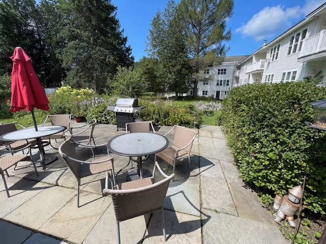 Outdoor patio at a senior living facility with round tables and wicker chairs, a red umbrella, a grill, and landscaped greenery in front of a white multi-story building.