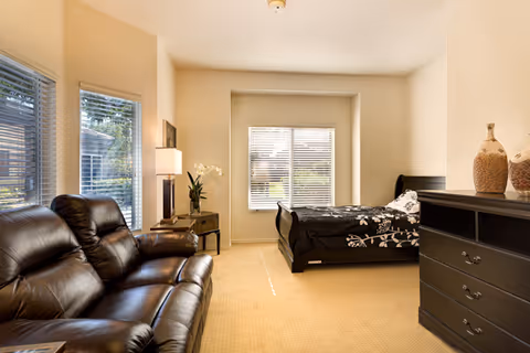 A well-lit bedroom featuring a black leather sofa on the left, a black bed with floral bedding near a window with blinds, a small side table with a lamp and a plant, and a black dresser with decorative vases on the right.