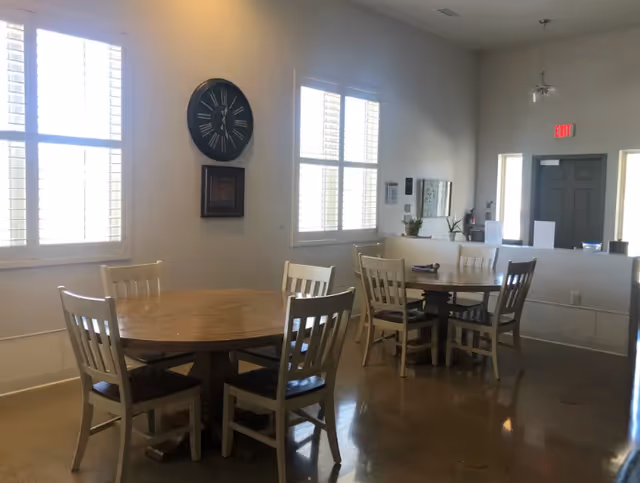 Interior view of a senior living facility dining area with two round wooden tables, each surrounded by four white wooden chairs. The room has large windows with white shutters allowing natural light to enter. A large black wall clock and a framed picture hang on the wall. The floor is polished concrete, and there is a door with an exit sign above it in the background.