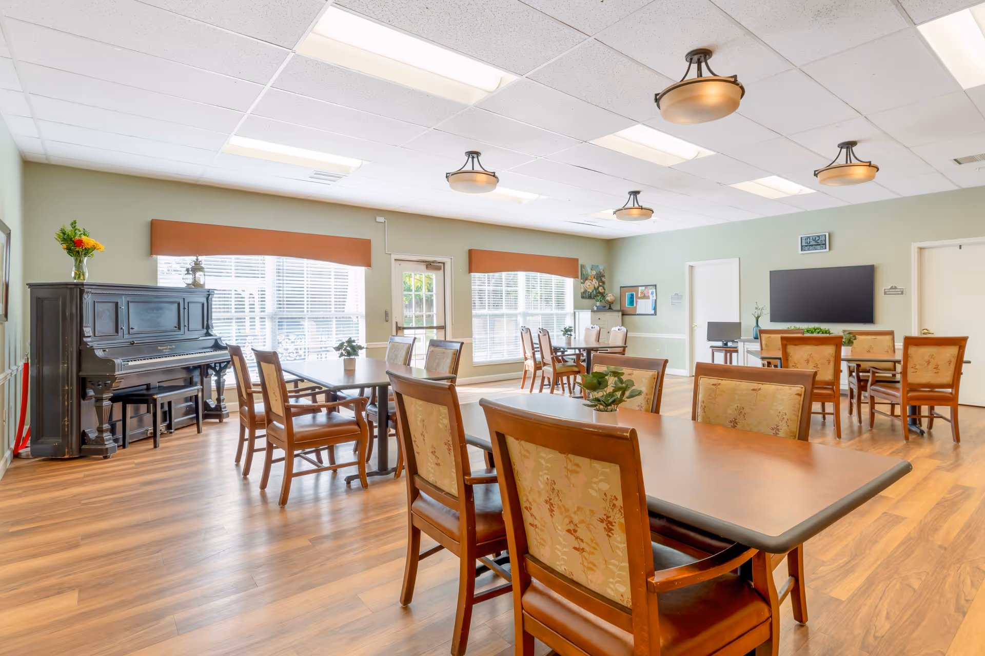 A bright and spacious common room with several wooden tables and chairs arranged neatly. The room features large windows with orange valances allowing natural light to fill the space. There is a black upright piano with a vase of flowers on top near the windows. The walls are painted light green, and the floor has wood-like laminate. Ceiling lights and a wall-mounted TV are visible, along with a clock and some decorative items on the walls.