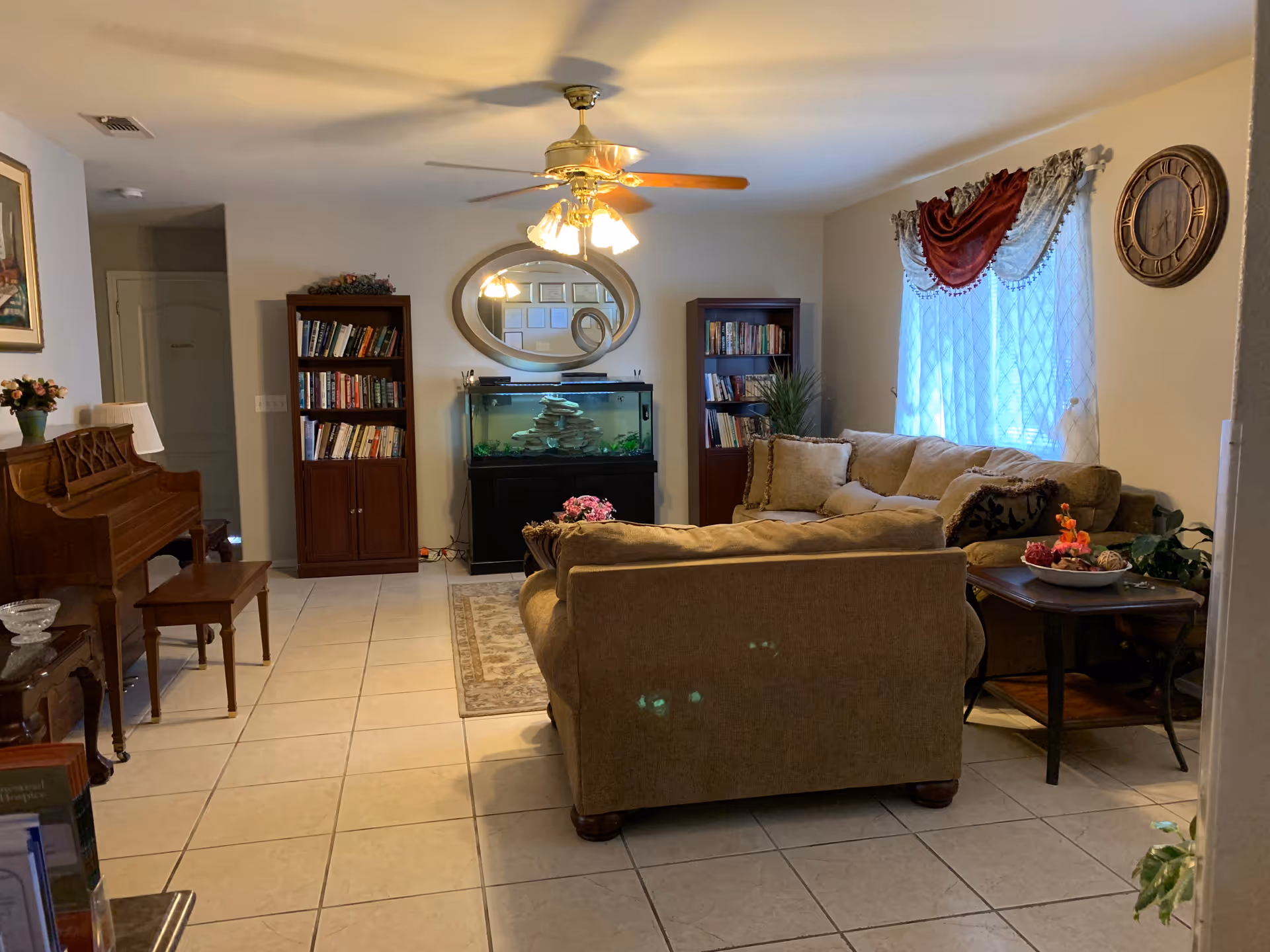 A cozy living room with beige tiled flooring, a ceiling fan with lights, a beige sofa set, a wooden piano with a bench, two wooden bookshelves filled with books, a large fish tank under an oval mirror, a side table with decorative items, a window with sheer white curtains and a red valance, and a wall clock.