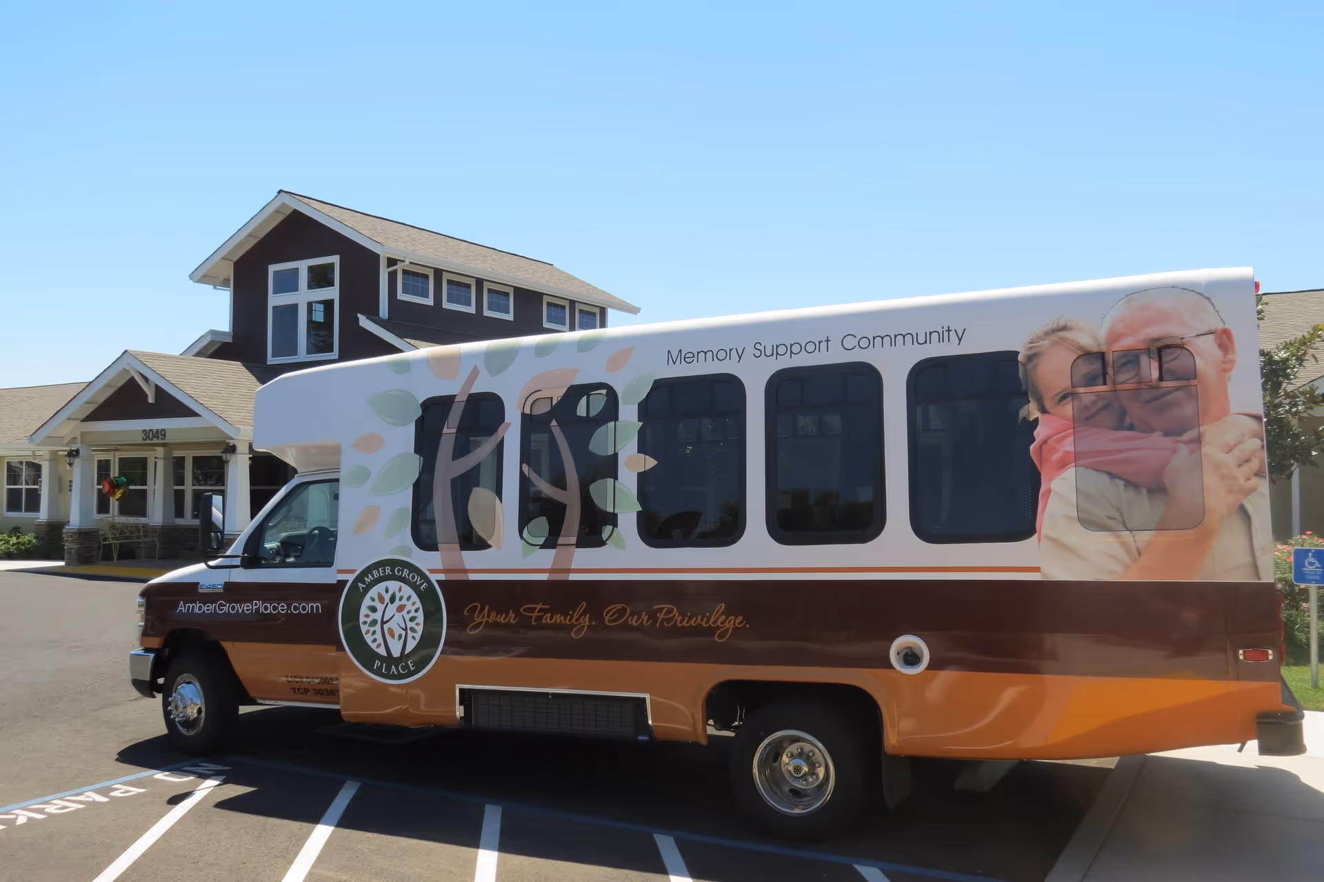 A shuttle bus parked outside a building with a sign reading 3049. The bus is branded with Amber Grove Place Memory Support Community logos and graphics, including an image of an elderly man hugging a young girl. The bus has the text 'Your Family. Our Privilege.' and the website AmberGrovePlace.com.