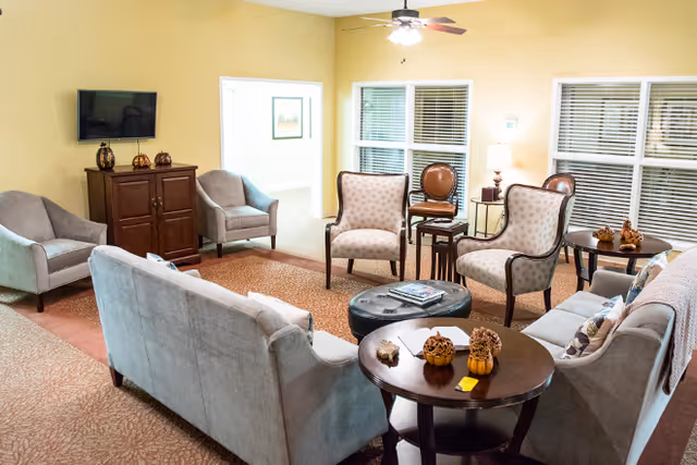 A cozy living room with beige walls and carpet, featuring a ceiling fan with light. The room contains multiple upholstered chairs and sofas arranged around a central ottoman and side tables decorated with small pumpkins and pinecones. A flat-screen TV is mounted on the wall above a wooden cabinet. Large windows with blinds allow natural light into the space.