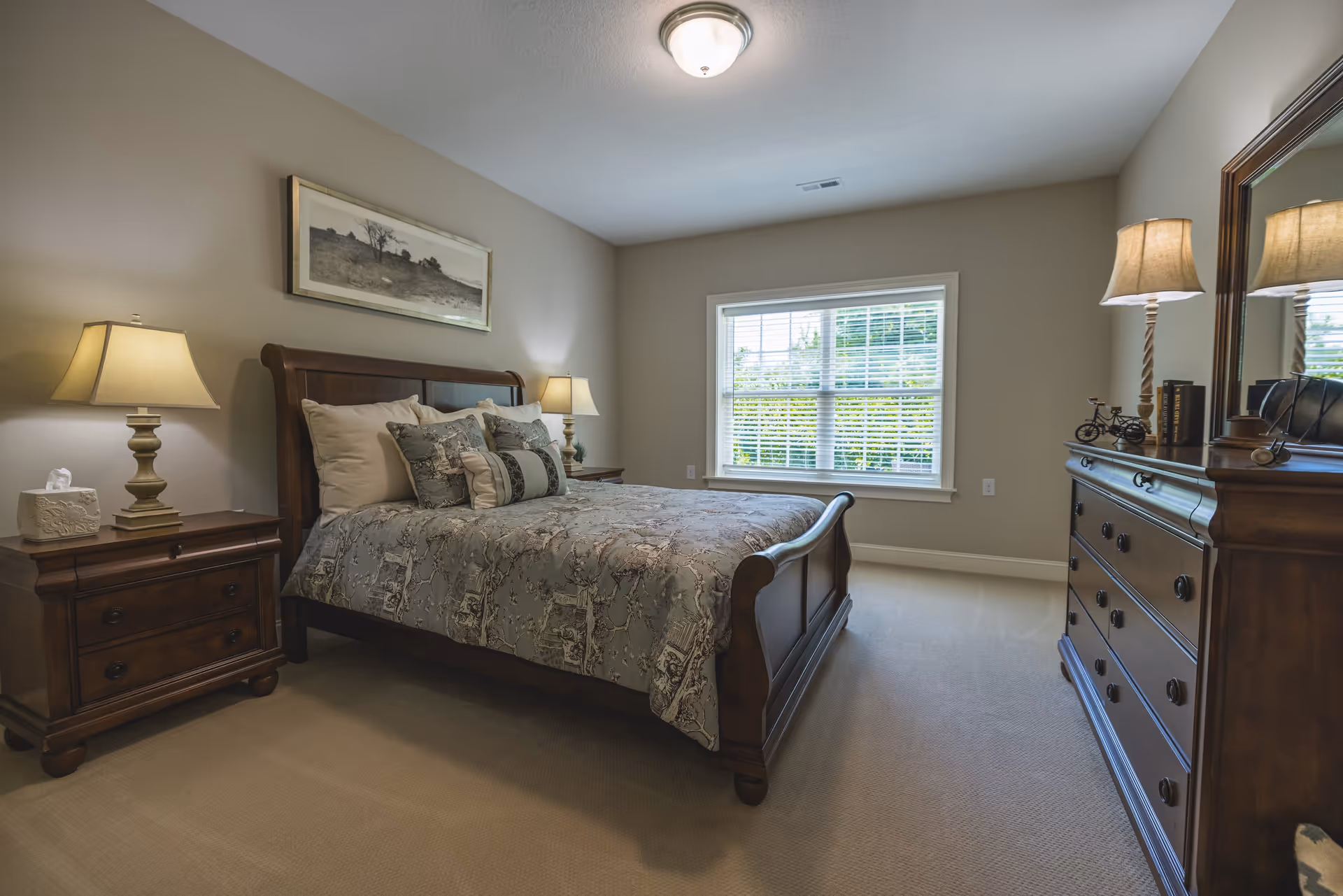 A neatly arranged bedroom with a wooden bed frame, patterned bedding, two bedside tables each with a lamp, a large window with white blinds, a wooden dresser with a mirror, and a framed picture on the wall.